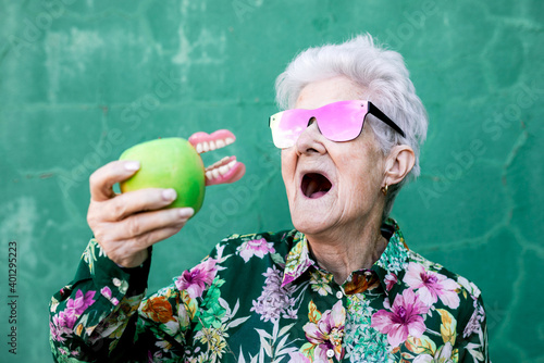 Stylish elderly female in trendy outfit and sunglasses holding green apple with denture while standing against green background photo