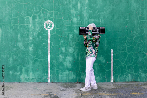 Full body side view of unrecognizable elderly gray haired female in stylish outfit carrying record player on shoulder while standing against green wall on street photo