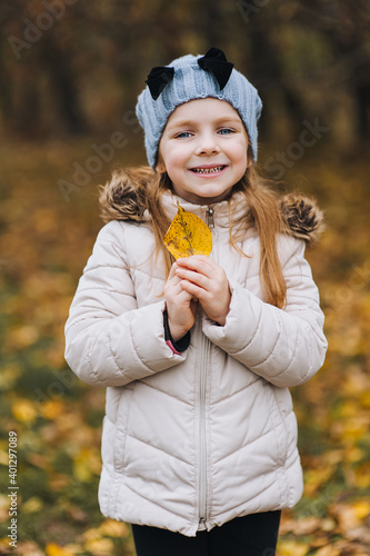 A beautiful cute girl  a child in a white jacket of preschool age  stands and poses with a yellow leaf in nature. Autumn portrait  photography.
