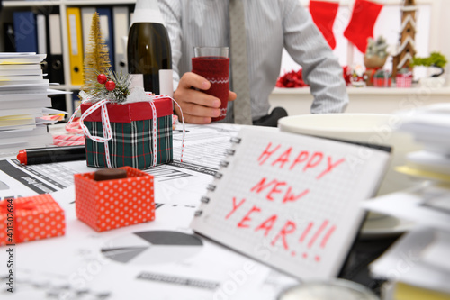 Business and holiday concept - businessman celebrating in office decorated with Christmas tree, candies and other new year decoration. Shows a notepad with happy new year text