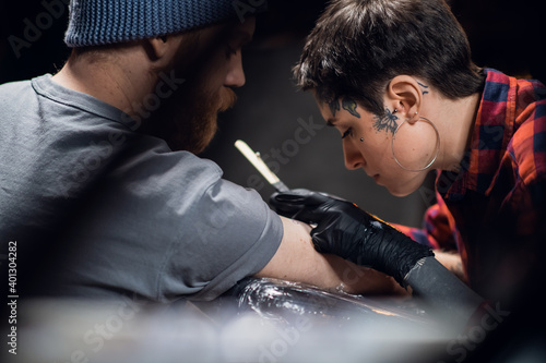 A young girl tattoo artist makes a bull tattoo on the arm of a young hipster man using the handpoke method in a tattoo parlor