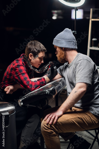 In the interior of a tattoo parlor, a young man with a beard is tattooed on his arm using the stick and poke method. Young artist girl in a checkered shirt