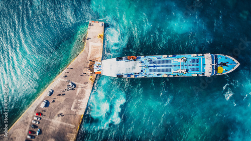 Aerial drone view of departing ferry at port with beautiful crystal blue water. Luxury cruise on a sunny day Greece. People and cars waiting in the harbour. Docking ship.