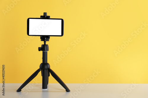 Smartphone with blank screen fixed to tripod on white table against yellow background. Space for text photo