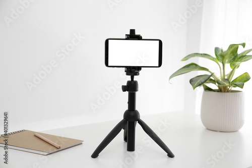 Smartphone with blank screen fixed to tripod on white table indoors. Mockup for design photo