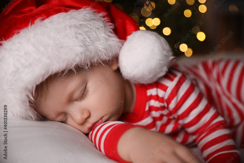 Baby in Christmas pajamas and Santa hat sleeping on bed indoors Baby in Christmas pajamas and Santa hat sleeping on bed indoors