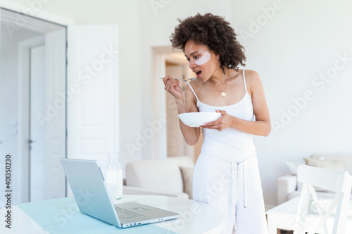 Beautiful young African American woman enjoys eating corn flakes for breakfast while working on her laptop in the morning. Looking at laptop computer and talking to her friends via video call. © Graphicroyalty