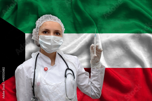 Young woman doctor in medical uniform on the background of the national flag of Kuwait is holding a syringe. The concept of a new vaccine against the disease, flu vaccine vaccination.