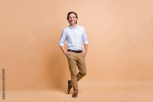 Full length body size view of representative cheerful man holding hands in pockets posing isolated over beige pastel color background