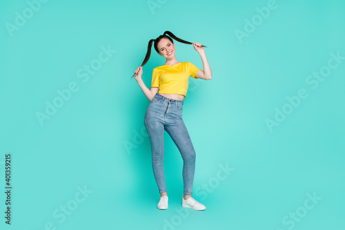 Full size photo of optimistic sweet brunette girl stand wear yellow t-shirt jeans sneakers isolated on cyan color background
