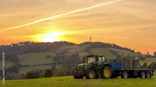 Timelapse close up of agricultural machinery tractor driving through a field and collecting bales of chopped hay on a hull during sunset over the horizon.