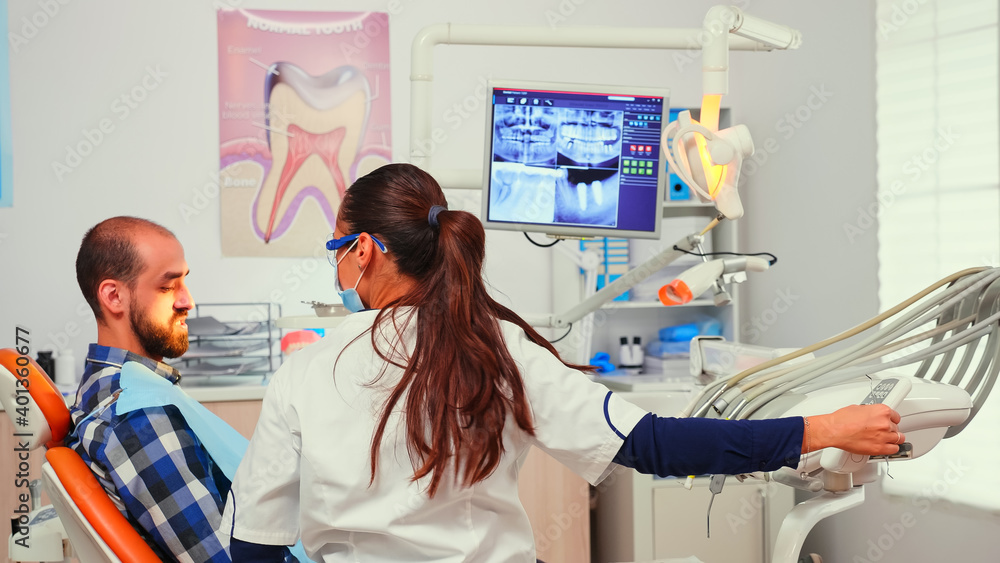 Obraz premium Stomatologist doctor lighting the lamp preparing patient for dental surgery. Dentist with face mask examining patient with dental bib before dental intervention sitting on stomatological chair