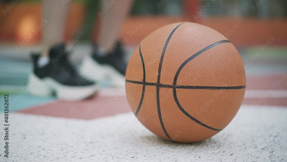 Close-up of orange ball with blurred sportswoman stretching feet at the background. Unrecognizable Caucasian basketball player warming up training outdoors. Workout and sport concept.