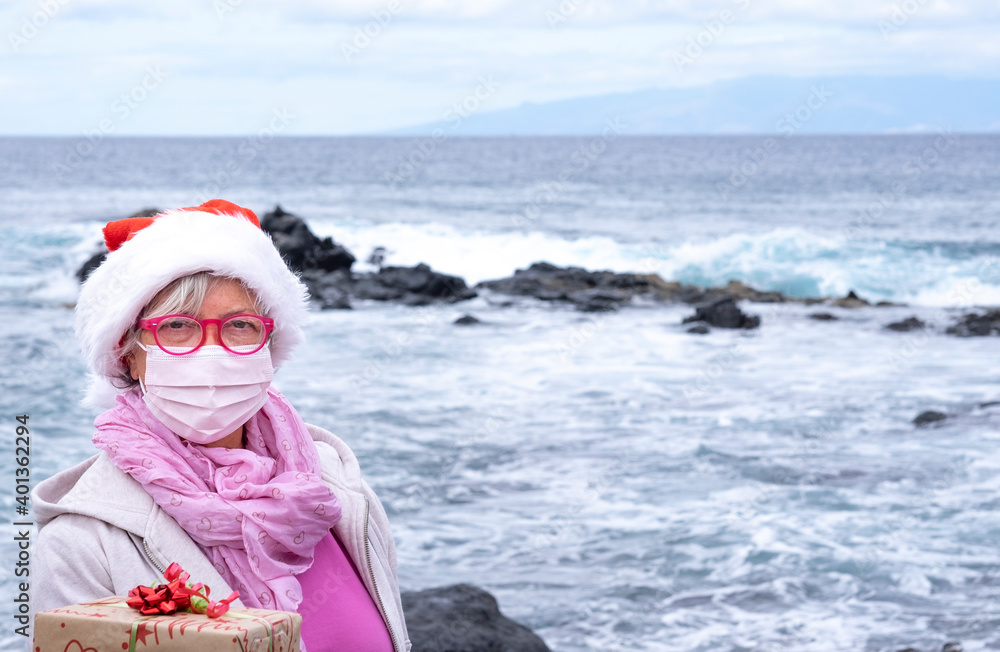 Obraz premium Senior woman in santa hat holding a christmas gift - wearing surgical mask due to coronavirus. Horizon over water