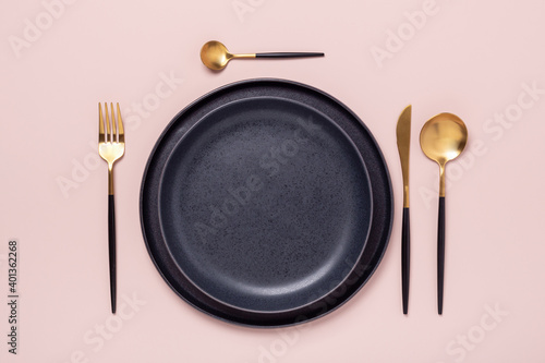 Dark ceramic plates and gold and black cutlery on pastel pink background. Top view