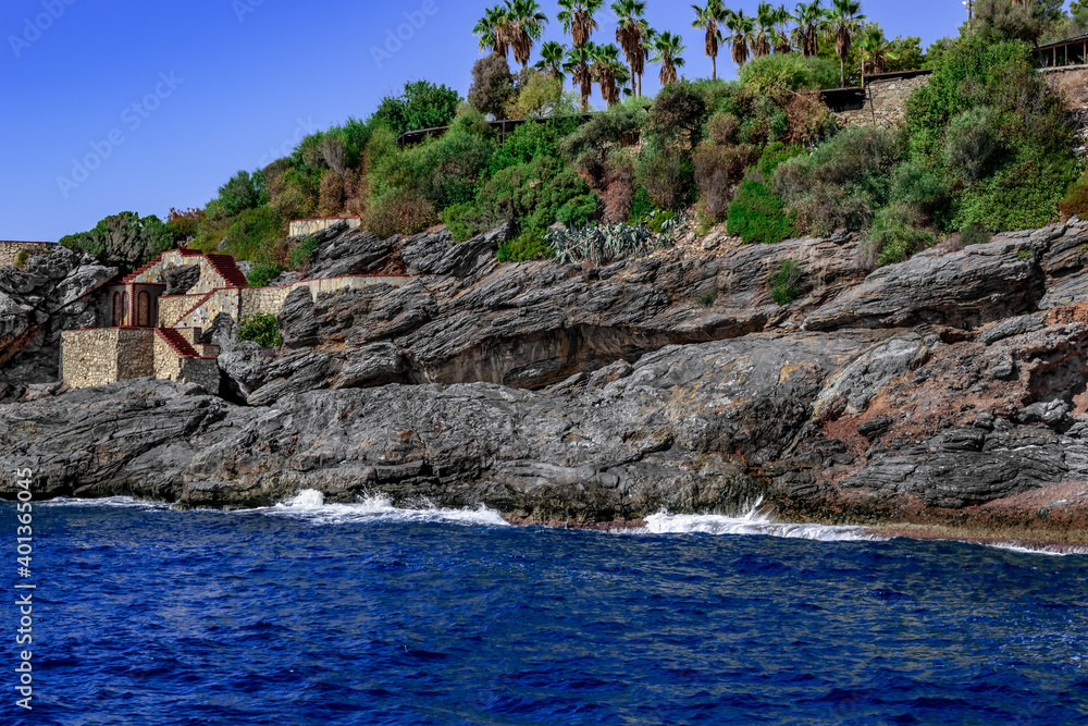 Layered granite cliff over the sea in the abandoned Emirgan Ulas Mesire Yeri Park in Alanya (Turkey). Stone steps in the rock against the background of blue water and tropical plants