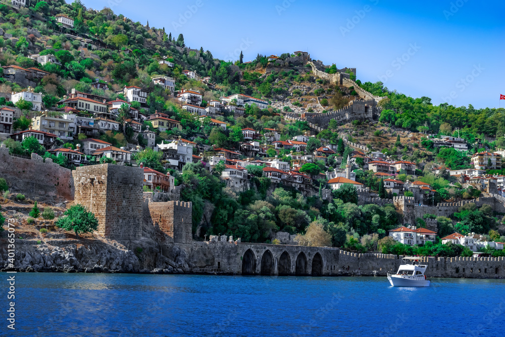 White boat in azure water against the background of an ancient arched wall and old town in Alanya (Turkey). Beautiful seascape with a historic resort city on the coast in the Turkish Riviera