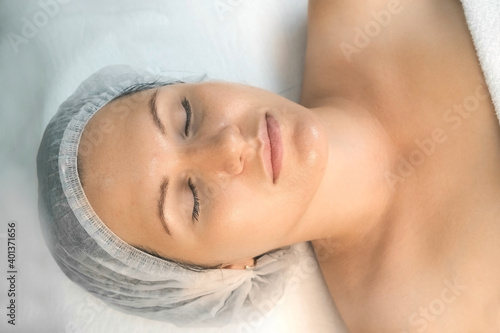 Woman on facial procedure in cosmetology clinic, closeup face, top view. She is lying on couch with closed eyes with yellow peeling on her face. Treatment, cure of face skin for young female.