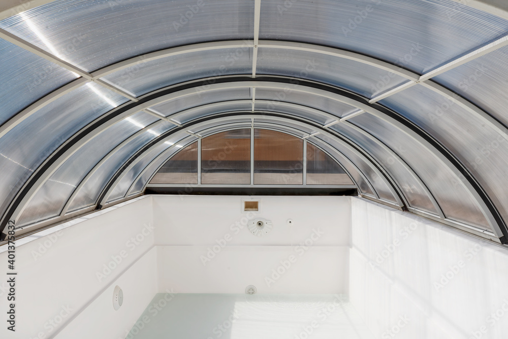 An empty fiberglass swimming pool with a sliding polycarbonate cover on a sunny spring day. A pool is white-colored, while the pavilion is half-transparent with blue toning. An empty fiberglass swimming pool with a sliding polycarbonate cover on a sunny spring day. A pool is white-colored, while the pavilion is half-transparent with blue toning.