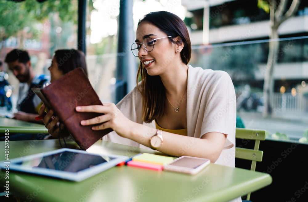 Fototapeta premium Happy female journalist checking informative notes in education textbook sitting in street cafe, successful hipster girl in optical eyewear for vision correction using personal planner outdoors