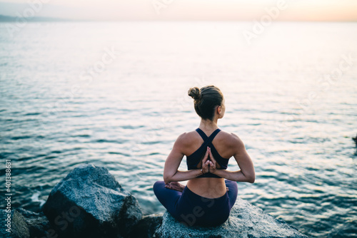 Back view of good looking female yogi practicing namaste breathing in Lotus pose recreating at seashore coastline, experienced fit girl in sportswear keeping body shape in tonus enjoying hatha