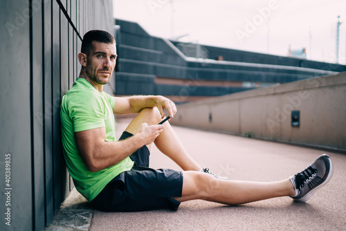 Portrait of muscular Caucasian athlete restine at urban setting of stadium and looking at camera during break time for smartphone networking, healthy sportsman holding cellular gadget in hand