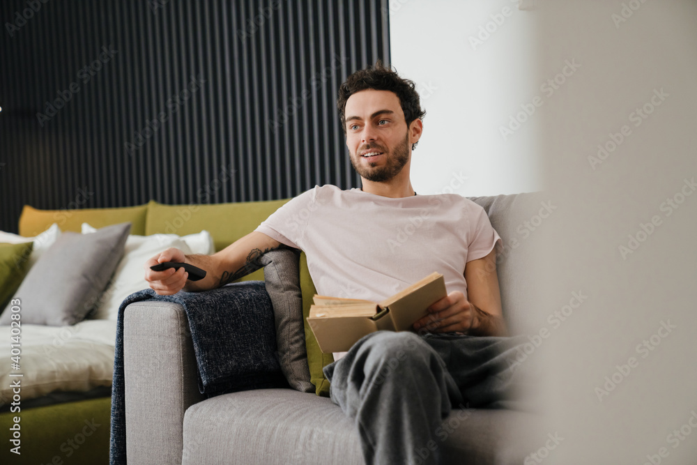 Pleased unshaven man using remote control while reading book on couch