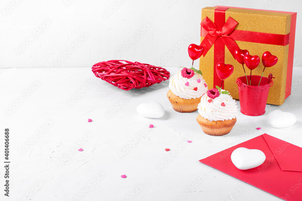 Valentines Day composition on the white table. Cupcakes with whipped cream and heart shaped sprinkles, red envelope, gift box and decorative hearts. Copy space Valentines Day composition on the white table. Cupcakes with whipped cream and heart shaped sprinkles, red envelope, gift box and decorative hearts. Copy space