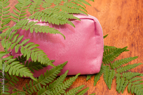 Bright pink cosmetic bag with spakles covered with green fern leaves on a wooden background photo