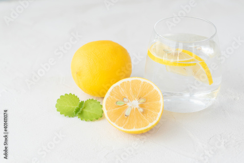 transparent glass with pure water, lemon slices, mint leaf ,  whole lemon and half of it lie on  light background. Healthy vitamin drink