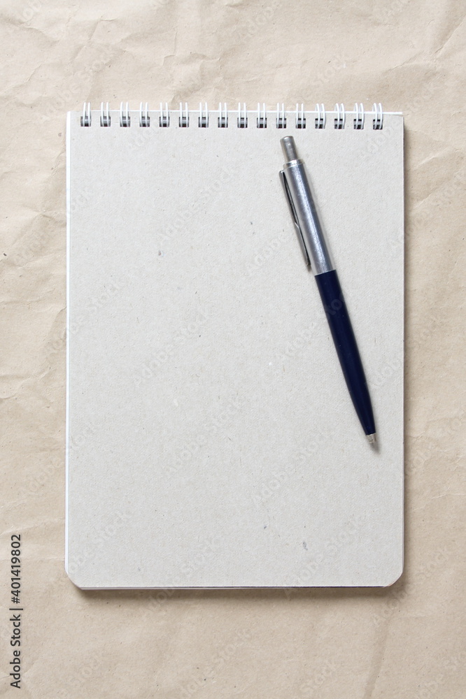 Gray notepad with white coiled spring and pen on a background of beige crumpled craft paper Gray notepad with white coiled spring and pen on a background of beige crumpled craft paper