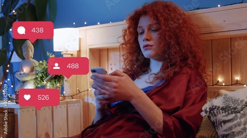 Attractive redheaded young relaxed woman lying in bed before sleep using smartphone interacting with social media folowers. Like icons. Communication. Home interior.