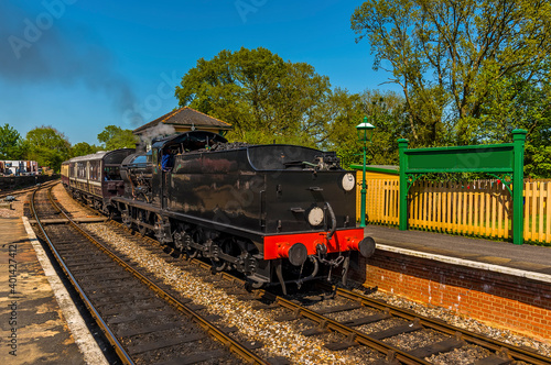 A steam train going past a station towards a single track section of line on a railway in the UK on a sunny summer day