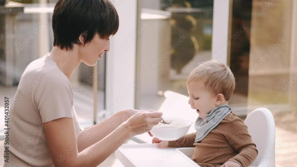 Mother feeding her baby son with spoon. Mother giving healthy food to her adorable child at home. Happy baby boy with bright eyes eating porridge. Concept baby food. Mother feeding her baby son with spoon. Mother giving healthy food to her adorable child at home. Happy baby boy with bright eyes eating porridge. Concept baby food.