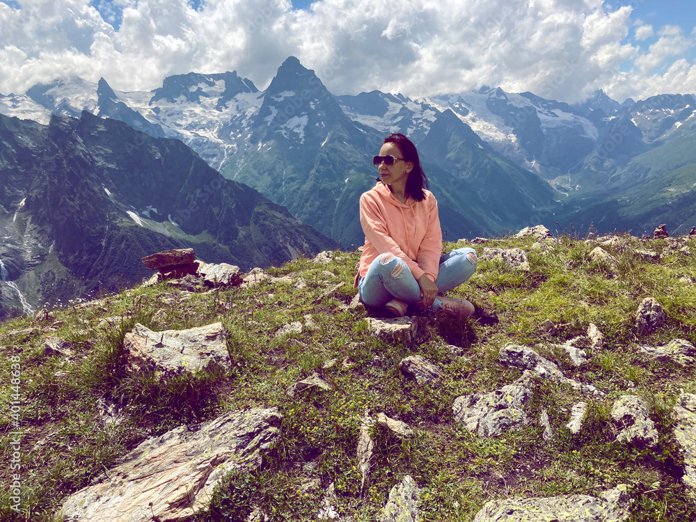 Young woman in sunglasses sitting on mountain top in sunny weather. Female tourist posing for camera on background of amazing mountain landscape in summertime.