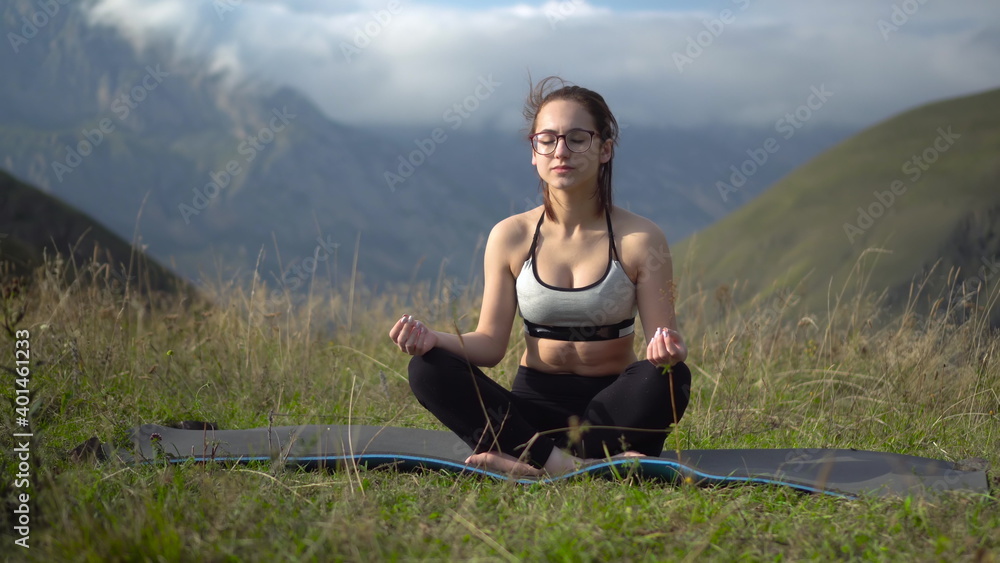 Young woman in tracksuit practices yoga performing lotus position in the mountains. The camera moves creating a parallax effect.