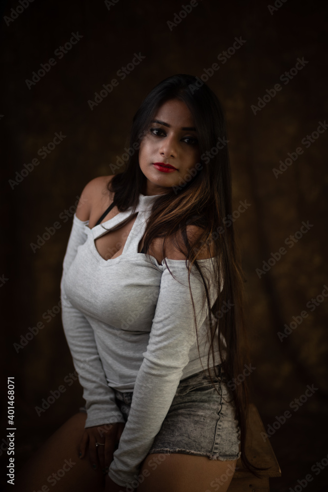 Studio portrait of a beautiful and young Indian Bengali female model in white cold shoulder top and jeans hot pant. Indian fashion and models
