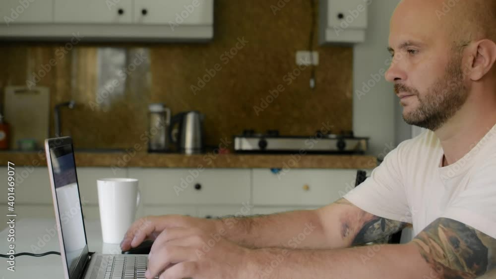 Tattooed man uses laptop while sitting at home in the kitchen. Tattooed man uses laptop while sitting at home in the kitchen.