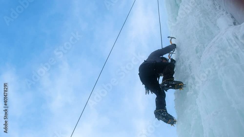 Alpinist man with ice tools axe climbing a large wall of ice.