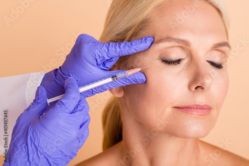 Close up photo middle aged woman undergo plastic surgery doctor make injection with syringe isolated beige color background photo