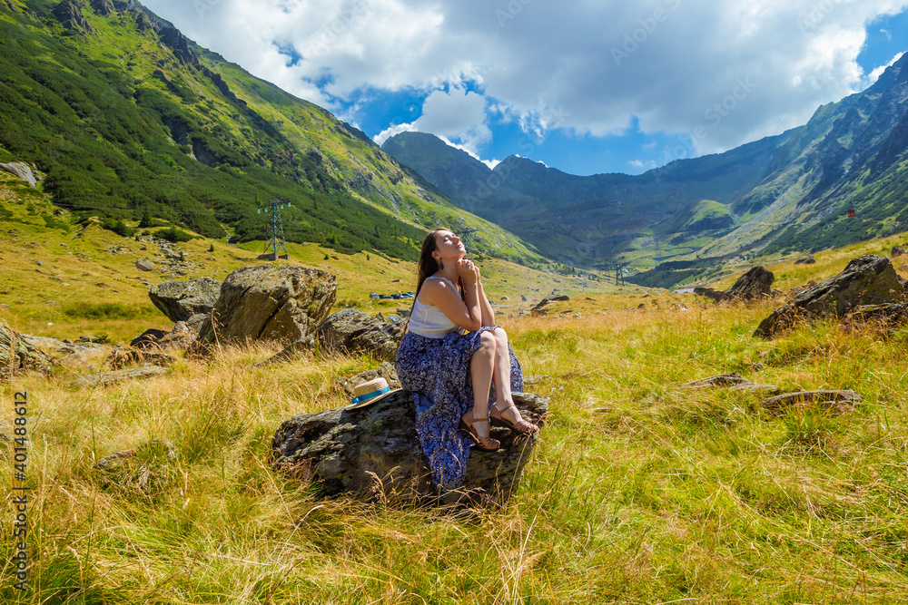Sensual pretty girl in long dress sits on a rock and dreaming against the romanian mountains background. Woman sitting near Transfegerasan, one of the most spectacular mountain road in the world