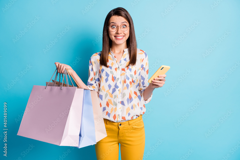 Photo of girl hold cellphone many packages toothy smile wear spectacles geometry print shirt yellow pants isolated blue color background Photo of girl hold cellphone many packages toothy smile wear spectacles geometry print shirt yellow pants isolated blue color background
