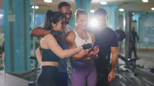 Multi-ethnic young foru frienda athletes standing together inside gym workout club using smartphone browsing online apps together. Friendship team. People in sports. photo