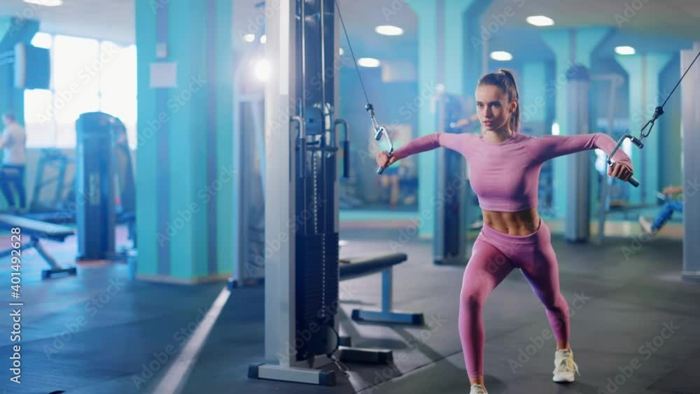 Side view of strong caucasian superwoman pulling power string weights machine in the gym. Female athlete exercising alone in sports club. Side view of strong caucasian superwoman pulling power string weights machine in the gym. Female athlete exercising alone in sports club.