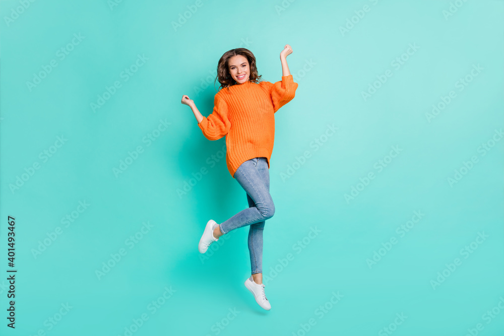 Full size photo of young happy positive excited girl jumping in victory wear orange pullover isolated on teal color background