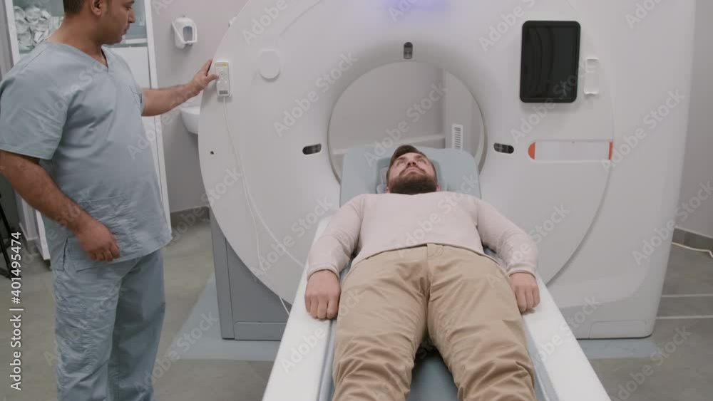 Handheld tracking of Arab male tech using control panel and moving patient lying on treatment couch into CT scan to perform procedure Handheld tracking of Arab male tech using control panel and moving patient lying on treatment couch into CT scan to perform procedure