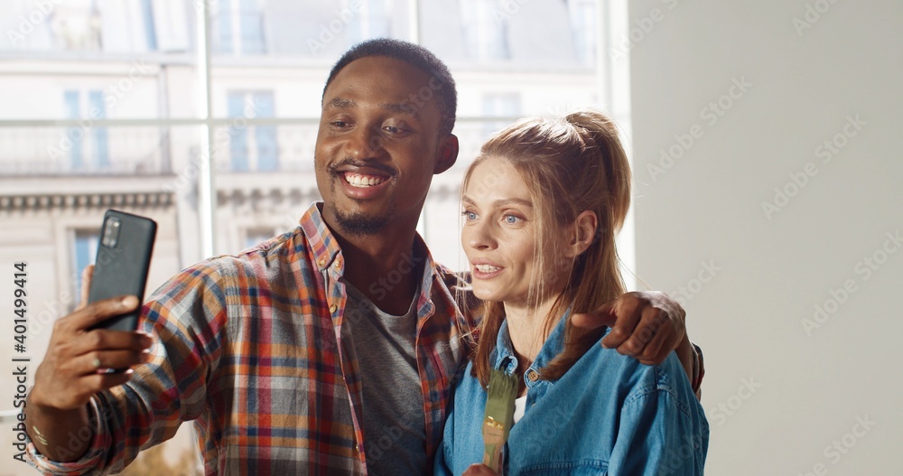 Happy mixed-races young married couple hugging smiling and posing with brushes taking selfie photos on cellphone in room. Home repair concept. Male and female friends taking pictures during renovation