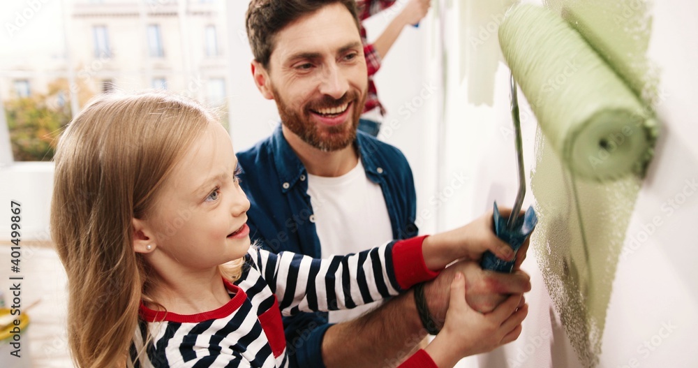 Portrait of cute family handsome happy father teaching little adorable kid girl daughter how to paint wall using paintbrush. Mother painting on background. Home renovation and repair concept Portrait of cute family handsome happy father teaching little adorable kid girl daughter how to paint wall using paintbrush. Mother painting on background. Home renovation and repair concept