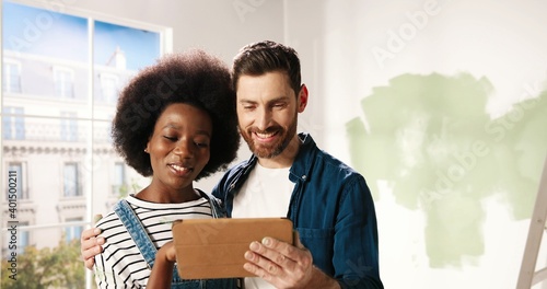 Fotografie Close up portrait of cheerful multi-ethnic young married couple Caucasian husband and African American wife standing in room typing on tablet device discussing design of new home