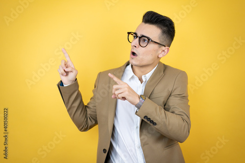 Young business man over isolated yellow background surprised and pointing his fingers side